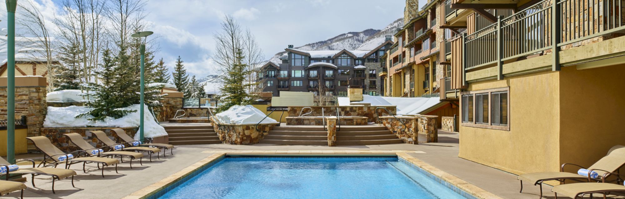 An outdoor pool area with lounge chairs, surrounded by snow, condo buildings, and trees; there's a mountain in the background.