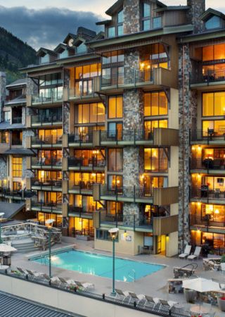 A multi-story hotel or condo building with numerous balconies, warmly lit windows, and a pool and patio area in the foreground, set against a mountain backdrop.