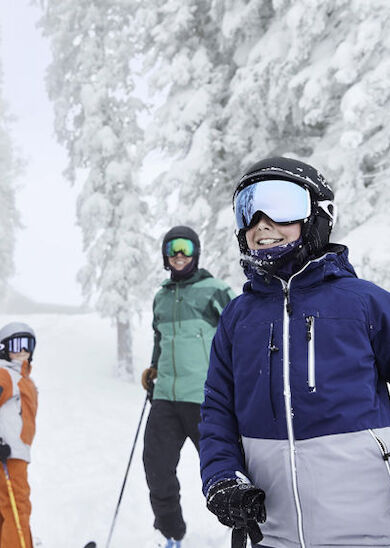 A group of three people in winter attire, including helmets and goggles, are skiing on a snow-covered landscape surrounded by snow-clad trees.