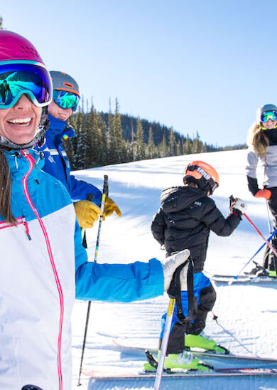 A group of people is skiing on a sunny day with snow-covered slopes and pine trees in the background.