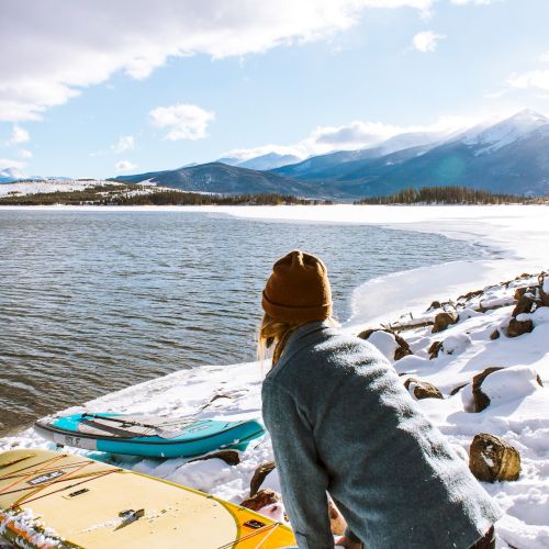 A person wearing a beanie and jacket is standing near paddleboards by a snow-covered lake with mountains in the background on a sunny day.