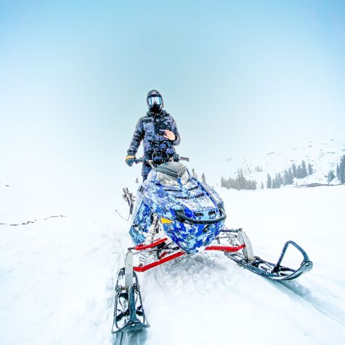 A person riding a snowmobile on a snowy landscape with a clear blue sky and distant trees in the background.