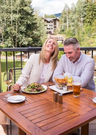 A smiling couple enjoying food and drinks at an outdoor restaurant with a scenic view of trees and buildings in the background.