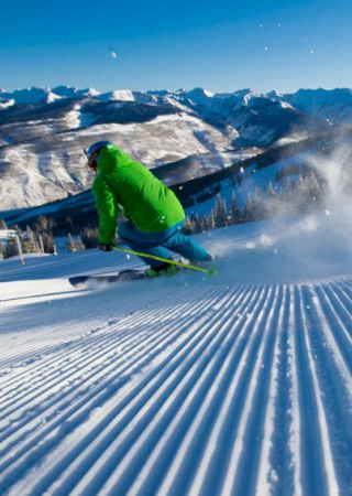 A skier in a green jacket descends a groomed slope with snow-capped mountains in the background under a clear blue sky.