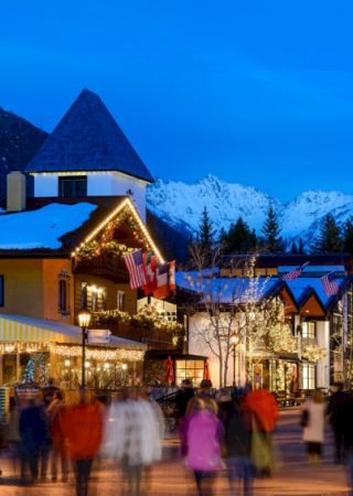 A lively town scene with people walking, featuring alpine-style buildings, colorful lights, and snow-covered mountains in the background.