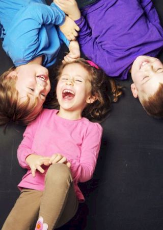 Three children are lying down and laughing, creating a playful and joyful atmosphere. They are dressed in colorful clothes on a dark surface.