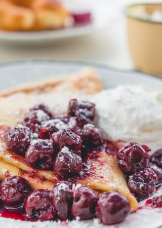 A plate of crepes topped with cherries and powdered sugar, served with a dollop of whipped cream, is shown in the foreground with coffee and pastries in the back.