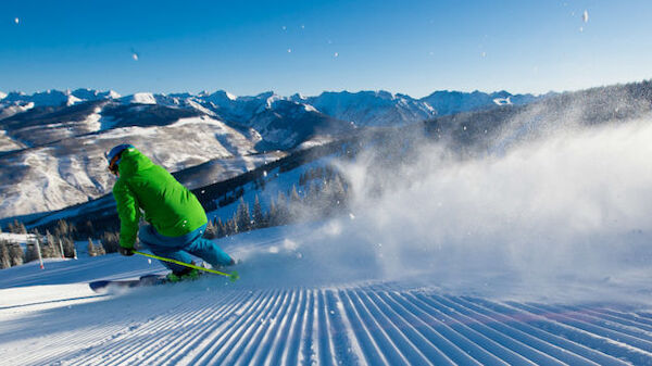 A skier in a green jacket is carving down a groomed slope, surrounded by mountains and a clear, blue sky.