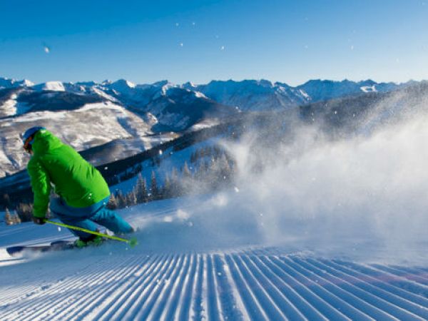 A skier in a green jacket is carving down a groomed slope, surrounded by mountains and a clear, blue sky.
