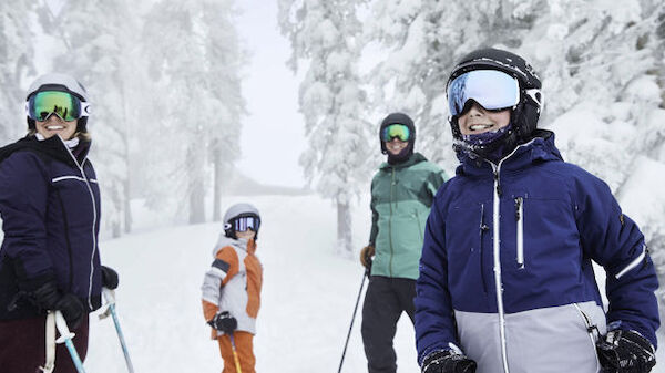 A group of four people, dressed in ski gear and helmets, stand on a snowy slope with trees covered in snow in the background.