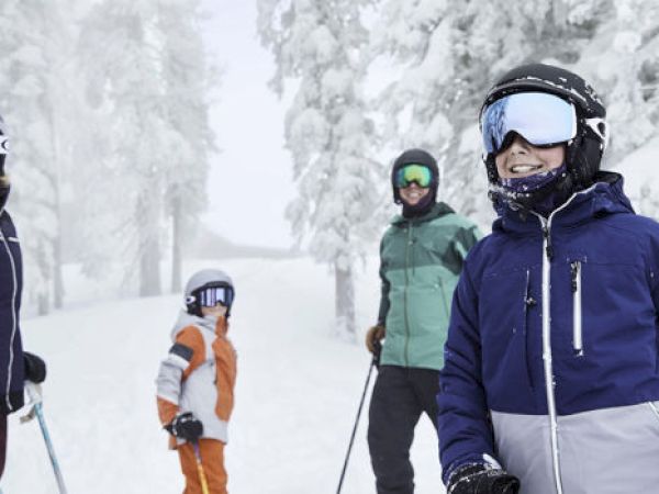 A group of four people, dressed in ski gear and helmets, stand on a snowy slope with trees covered in snow in the background.