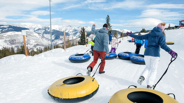 People are pulling snow tubes uphill in a snowy mountain landscape. The sky is clear with scattered clouds, and trees are in the background.