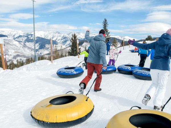 People are pulling snow tubes uphill in a snowy mountain landscape. The sky is clear with scattered clouds, and trees are in the background.