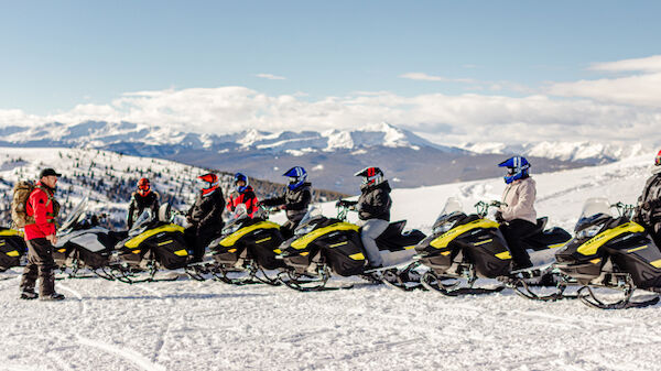 A group of people on snowmobiles on a snowy mountain, with a person in red standing nearby and mountains in the background.