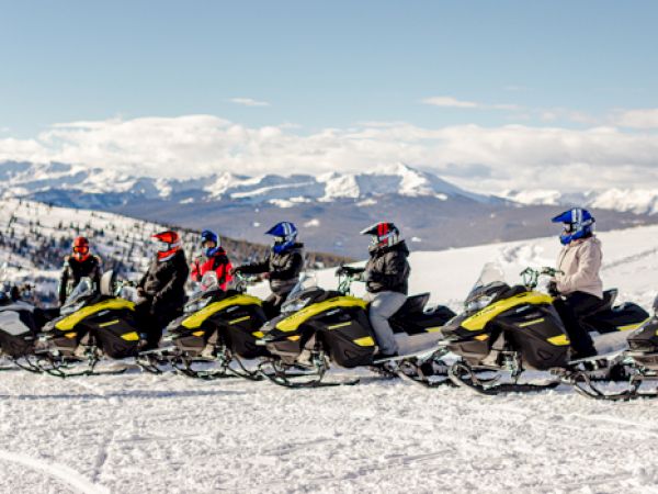 A group of people on snowmobiles on a snowy mountain, with a person in red standing nearby and mountains in the background.