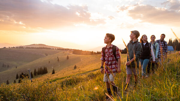 A group of people walks along a grassy hillside at sunset, with beautiful mountainous scenery in the background, enjoying an outdoor adventure.