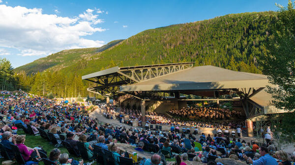 A large outdoor concert with a covered stage set amidst a scenic, mountainous backdrop. The seated audience enjoys the performance under a clear sky.