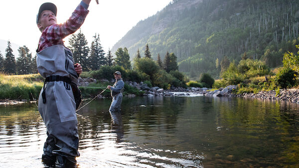 Two people fishing in a clear river surrounded by lush green mountains and trees, with one in the foreground casting their line.