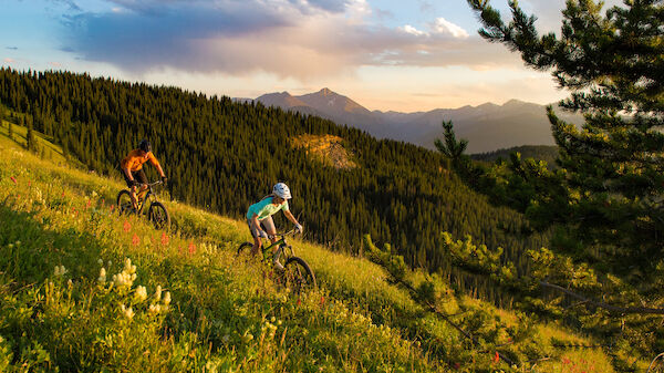 Two cyclists are riding mountain bikes on a grassy slope with scenic mountainous terrain in the background, under a partially cloudy sky.