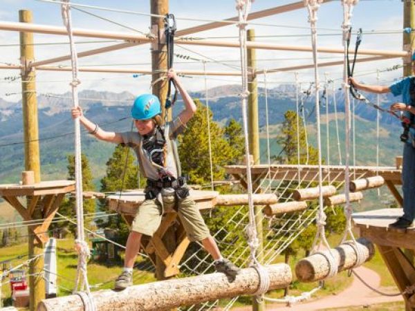 Two children are navigating a high ropes course, wearing helmets and safety harnesses, with a mountainous landscape in the background.
