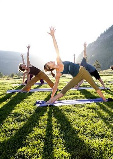 A group of people is practicing yoga outdoors on a grassy field with mountains and trees in the background, all stretching in a triangle pose.