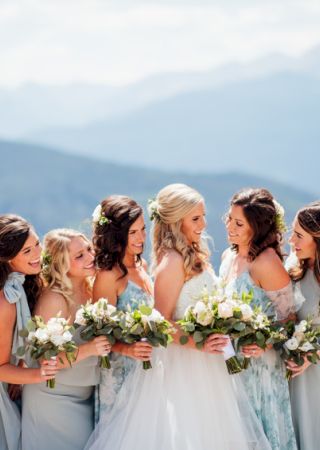 A bride and bridesmaids in pastel dresses are posing with bouquets, smiling against a mountain backdrop, creating a joyful wedding scene.