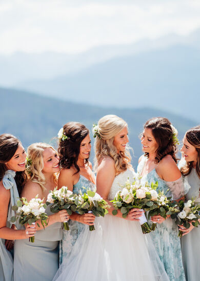 A bride and bridesmaids in pastel dresses are posing with bouquets, smiling against a mountain backdrop, creating a joyful wedding scene.