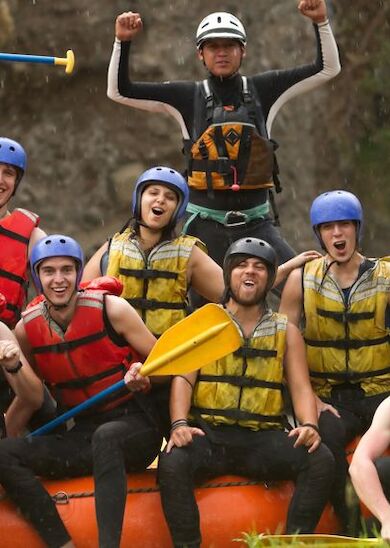 A group of people in helmets and life jackets are excitedly posing on a raft with paddles, showing thumbs up and celebratory gestures.