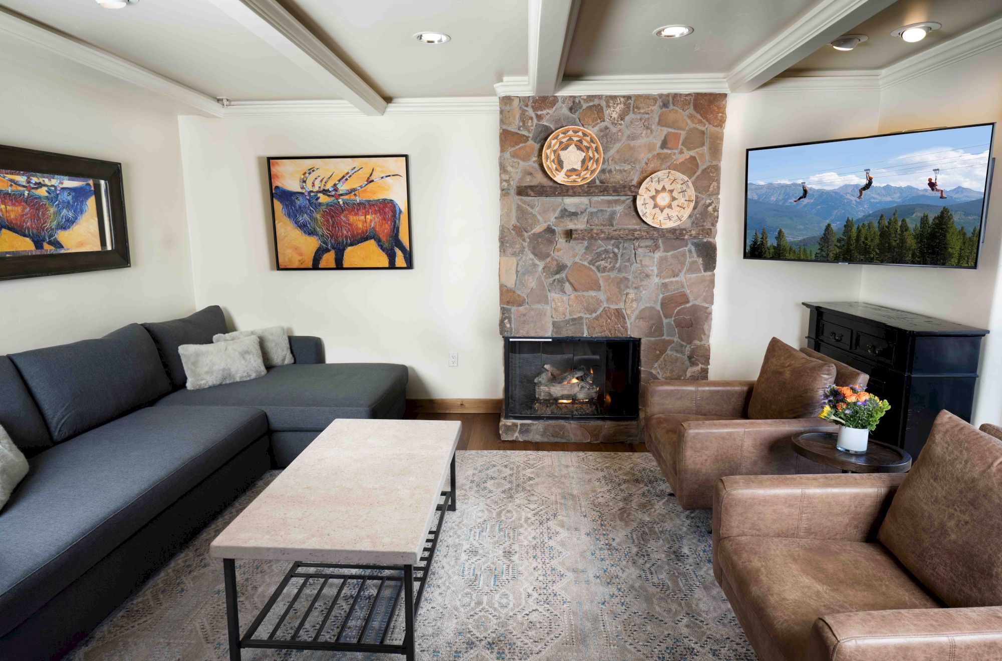 A cozy living room with a gray sectional sofa, two brown armchairs, a stone fireplace, artwork on walls, a coffee table, and a TV.
