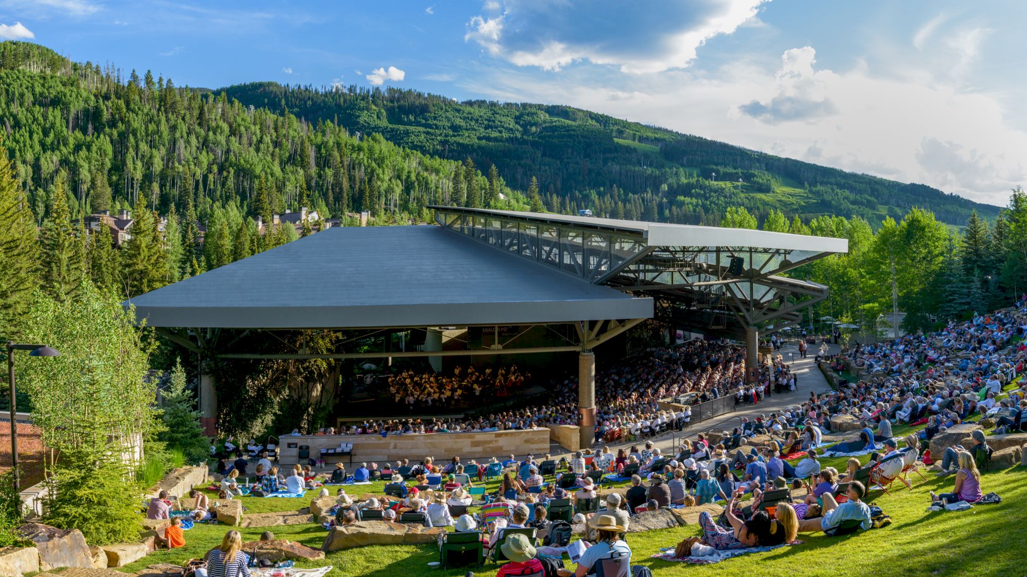 An outdoor amphitheater with people seated on a grassy slope, surrounded by lush greenery and mountains, under a partly cloudy sky.
