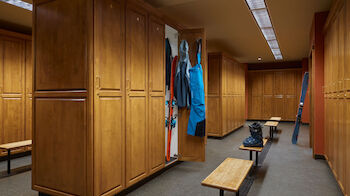 A spacious locker room with wooden lockers, benches, and ski gear hanging. The floor is clean, and the ceiling lights are bright.