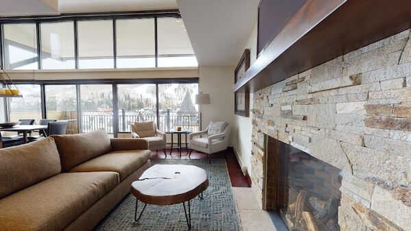 A cozy living room with a brown sofa, stone fireplace, large windows, and a round coffee table. It overlooks a snowy outdoor scene through the windows.