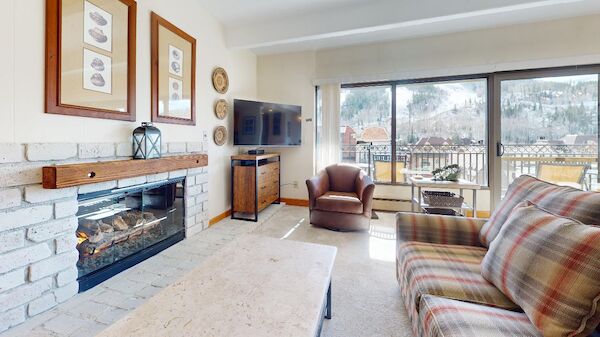 A cozy living room with a brick fireplace, TV, armchair, and plaid sofa. Large windows offer views of a snowy landscape outside.