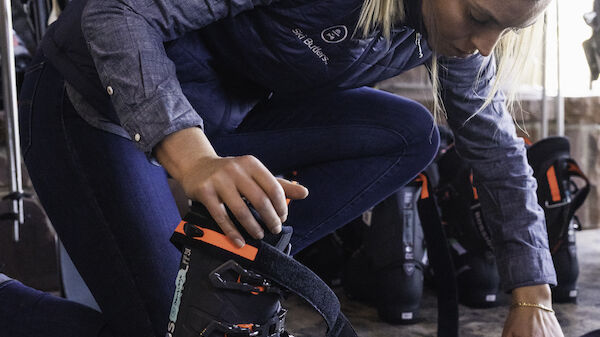 A person is adjusting ski gear indoors, possibly preparing for skiing, with other ski equipment visible in the background, wearing a grey jacket.