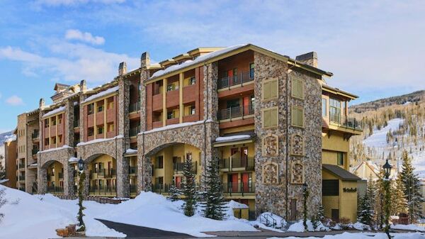 The image shows a multi-story building with a rustic design, surrounded by snow-covered ground and trees, set against a mountainous backdrop.