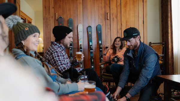 A group of people dressed warmly, some holding drinks, in a room with skis in the background. One person is tying another's shoe or adjusting gear.