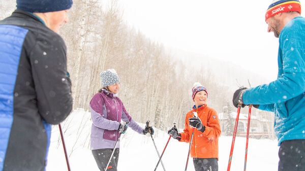 Four people dressed in winter gear are standing and talking while holding ski poles in a snowy outdoor setting.