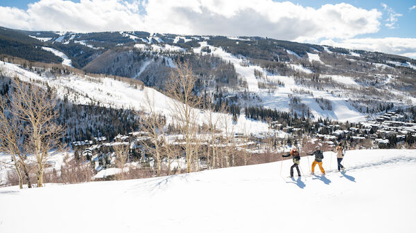 Three people are skiing on a snowy mountain with a scenic view of slopes, trees, and a town below under a partly cloudy blue sky.