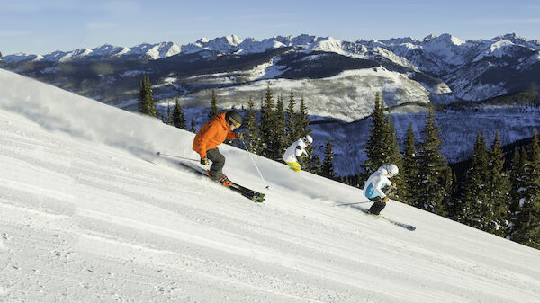 Three skiers are descending a snowy slope with a backdrop of pine trees and snow-capped mountains under a clear blue sky.