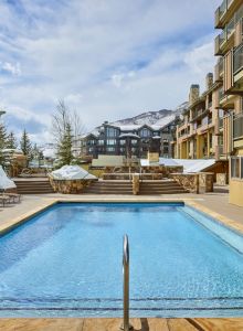 An outdoor swimming pool surrounded by lounge chairs, with a view of snowy mountains and residential buildings.