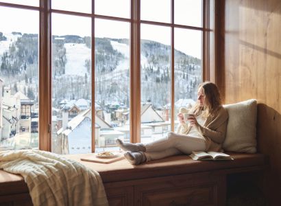 A person is sitting on a window seat, holding a cup, with a book beside them, overlooking a snowy mountain village through large windows.