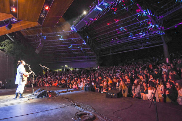 A musician performs on stage with a guitar, while a large, seated audience watches under a covered venue with colorful lights.